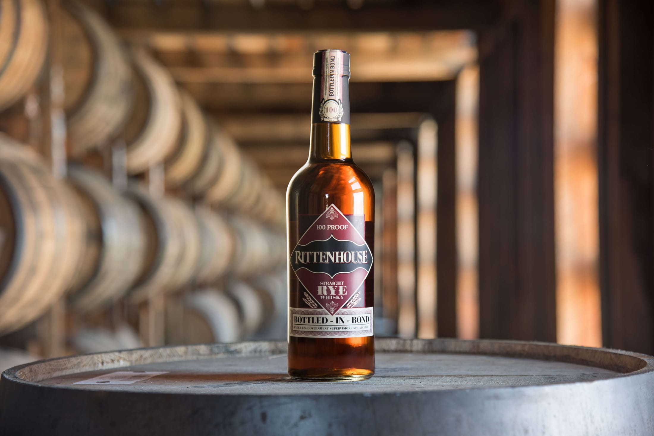Rittenhouse Bottled-in-Bond Straight Rye Whiskey Photocredit: Heaven Hill - a Bottle of Rittenhouse Rye sitting on a barrel in front of more aging barrels of rye whiskey